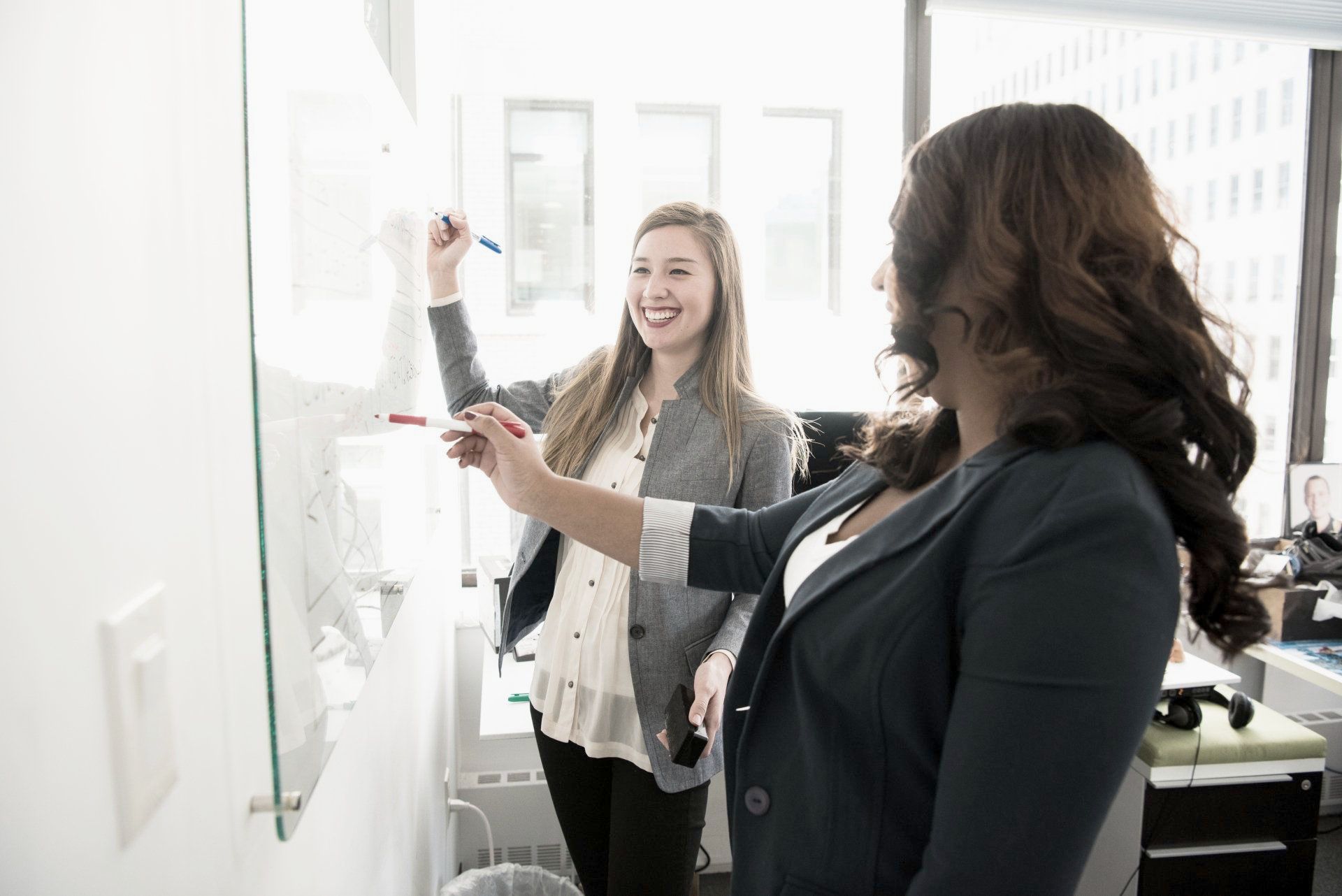 Two women are standing next to each other in front of a whiteboard.