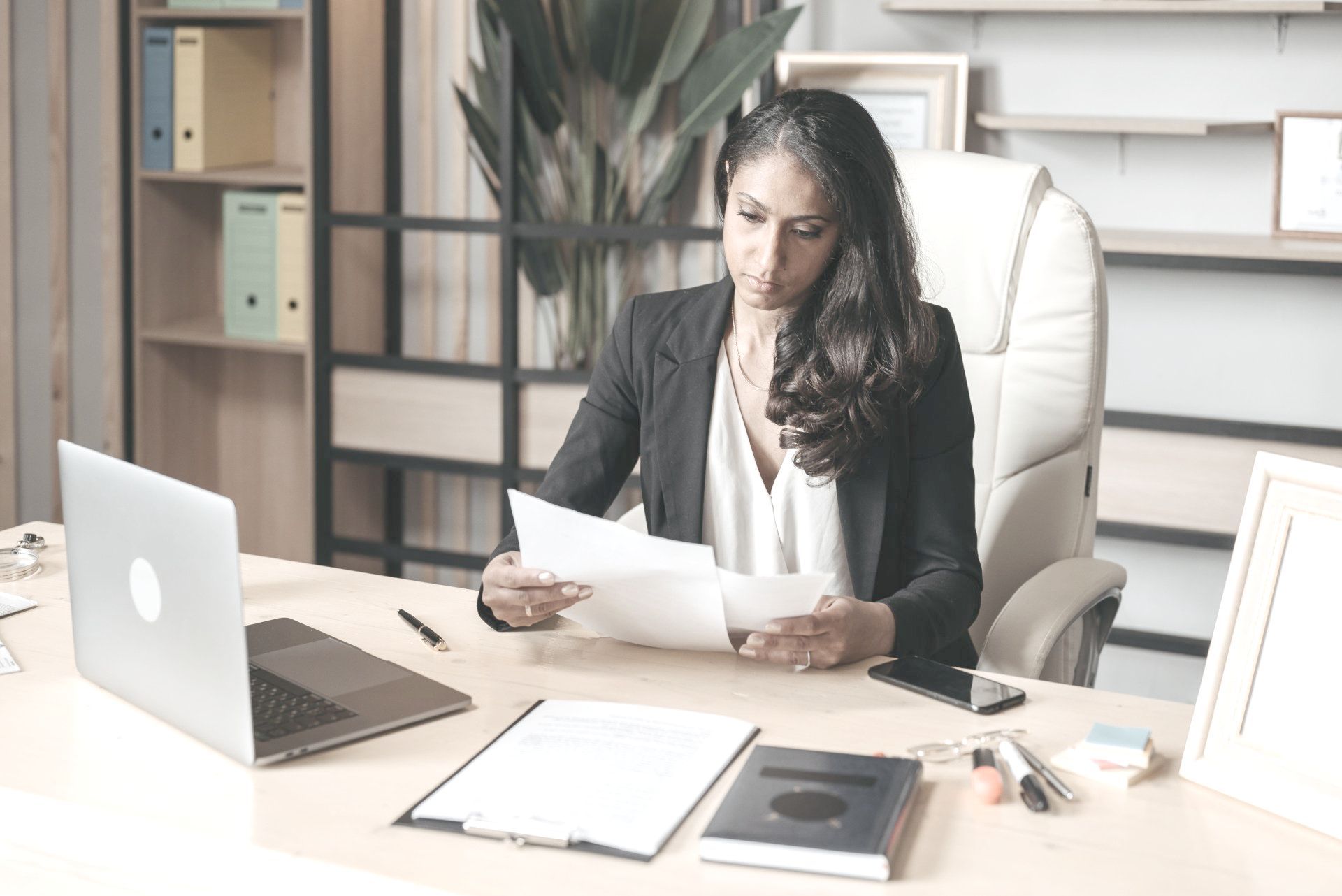 A woman is sitting at a desk with a laptop and a piece of paper.