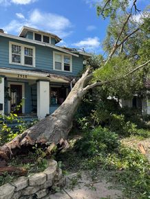 Storm-damaged house with a large fallen tree leaning across the yard and roof