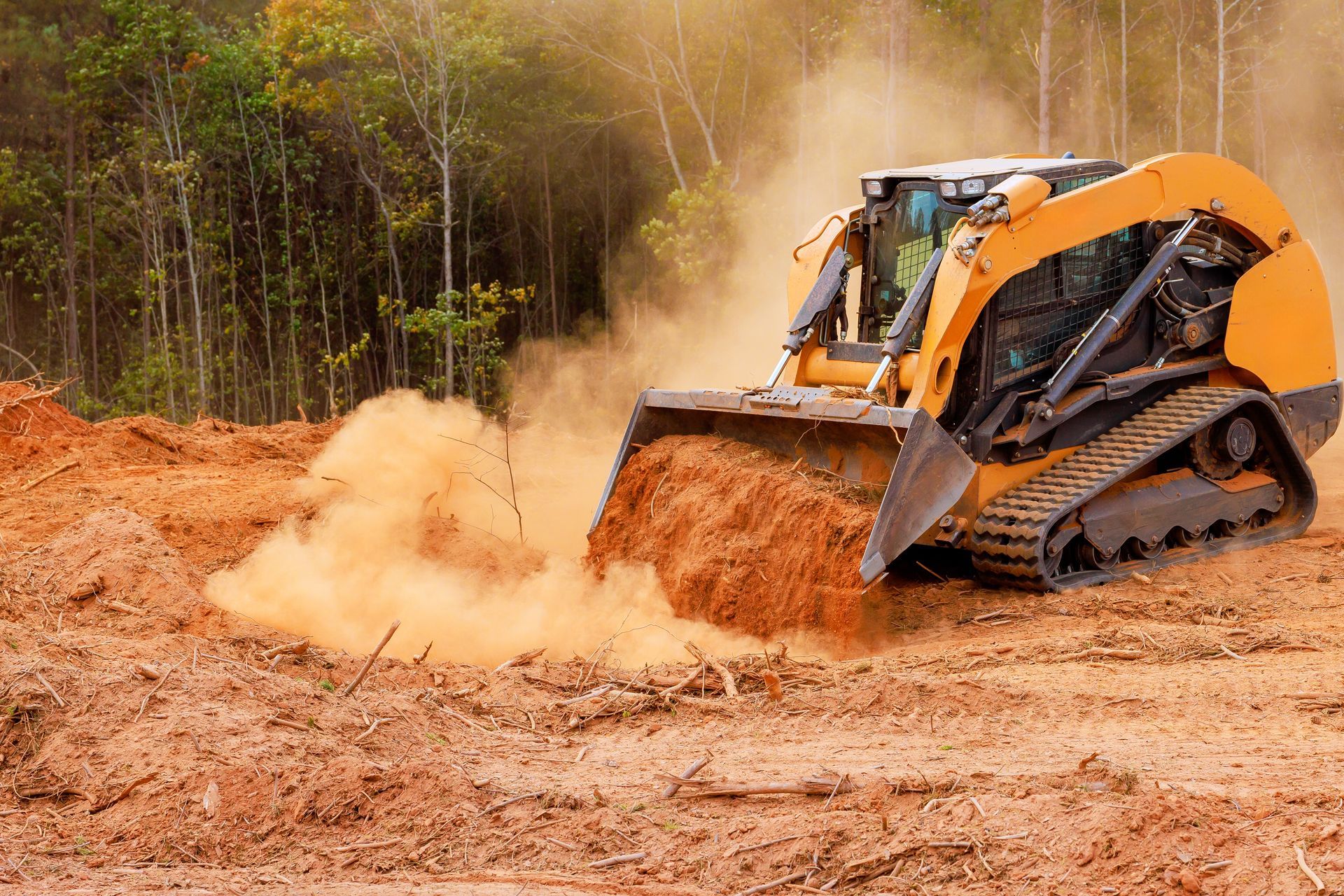 Orange skid-steer loader pushing dirt on a dusty construction site near a wooded area