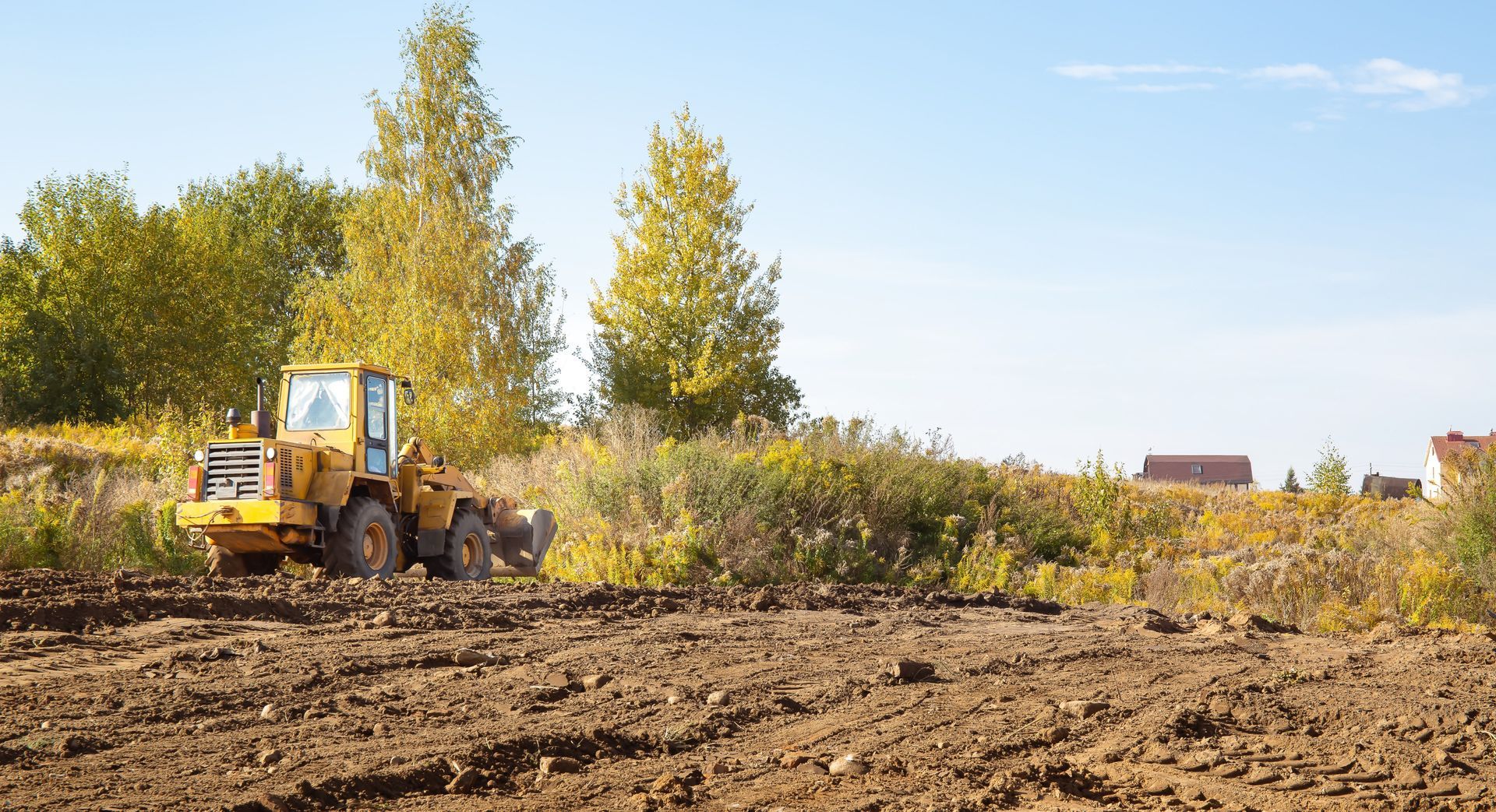 Yellow bulldozer grading a dirt field with trees and blue sky in the background