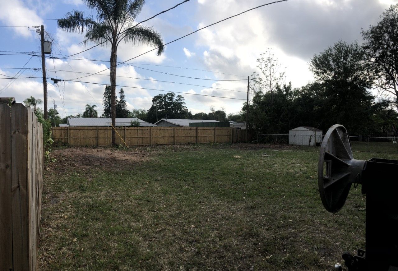 Backyard with patchy grass, palm trees, a wooden fence, and cloudy sky; black grill on the right.