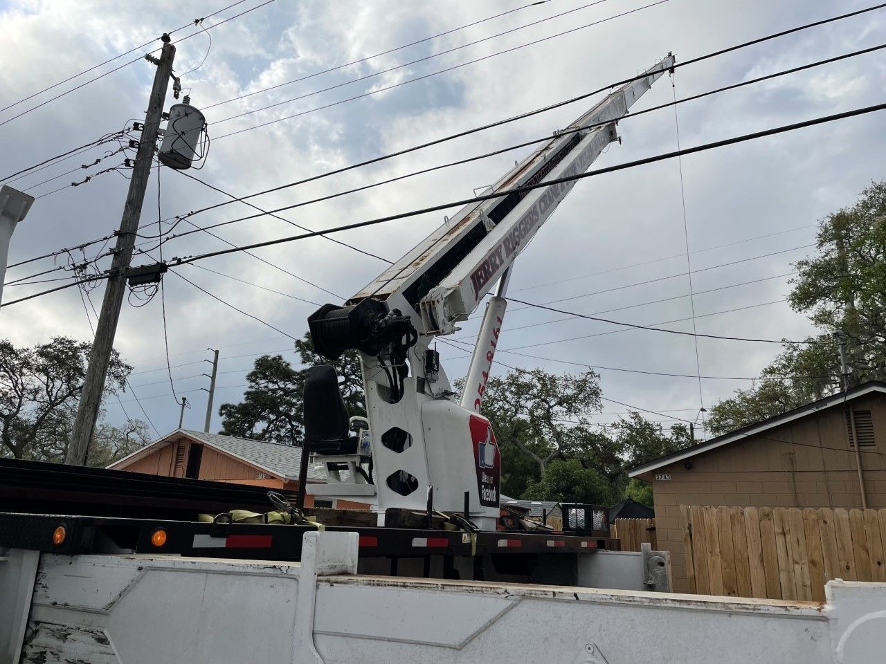 Tow truck with raised boom parked by a street under cloudy sky, near houses and power lines