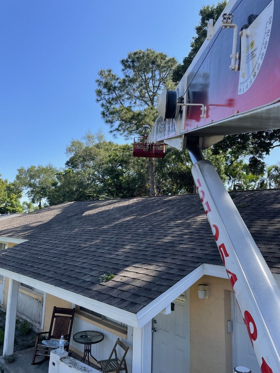 Worker in a bucket lift beside a house roof, trimming a tree under a clear blue sky.