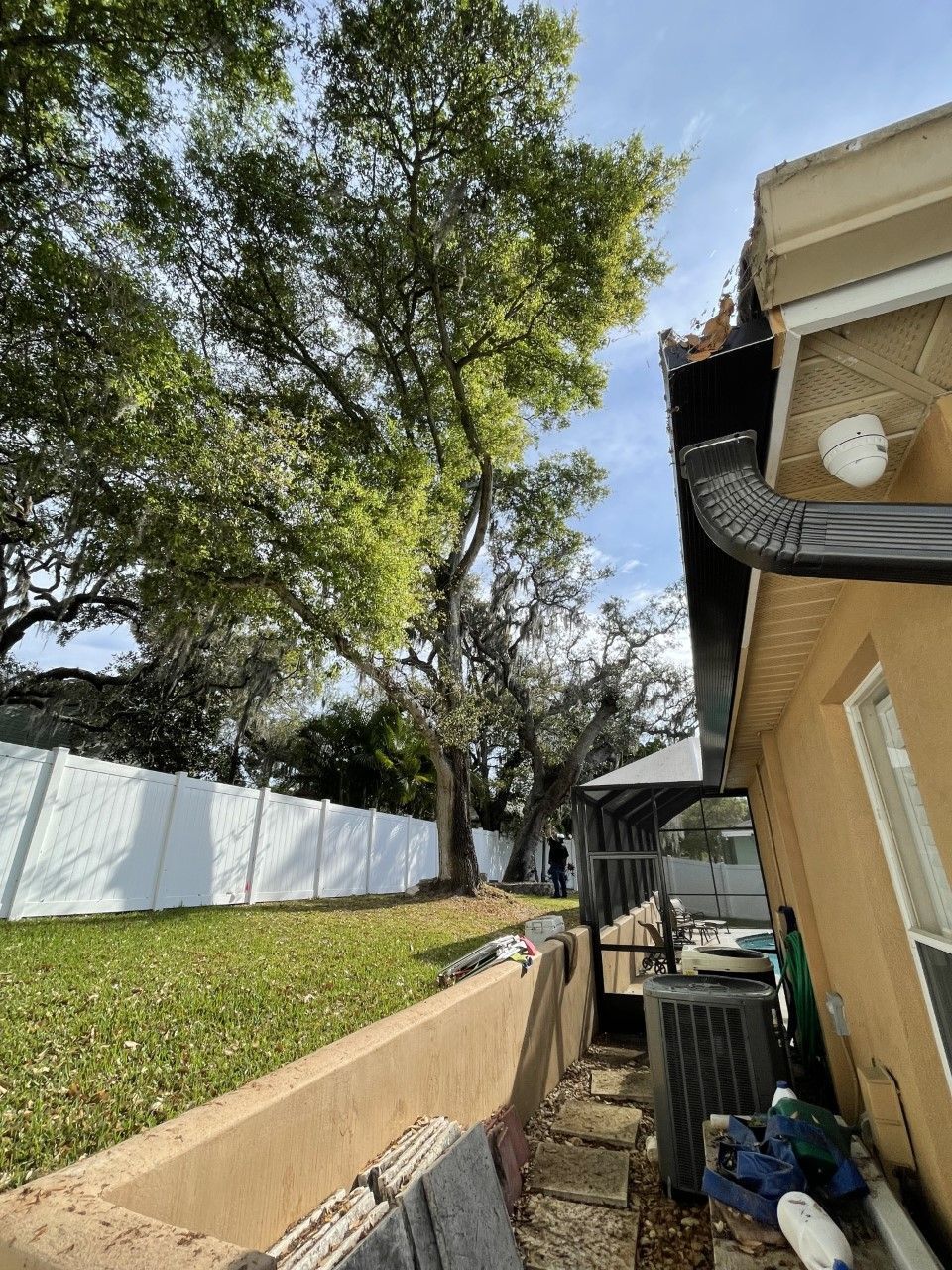 Backyard with large tree, white fence, tan house, and drainage trench beside the wall