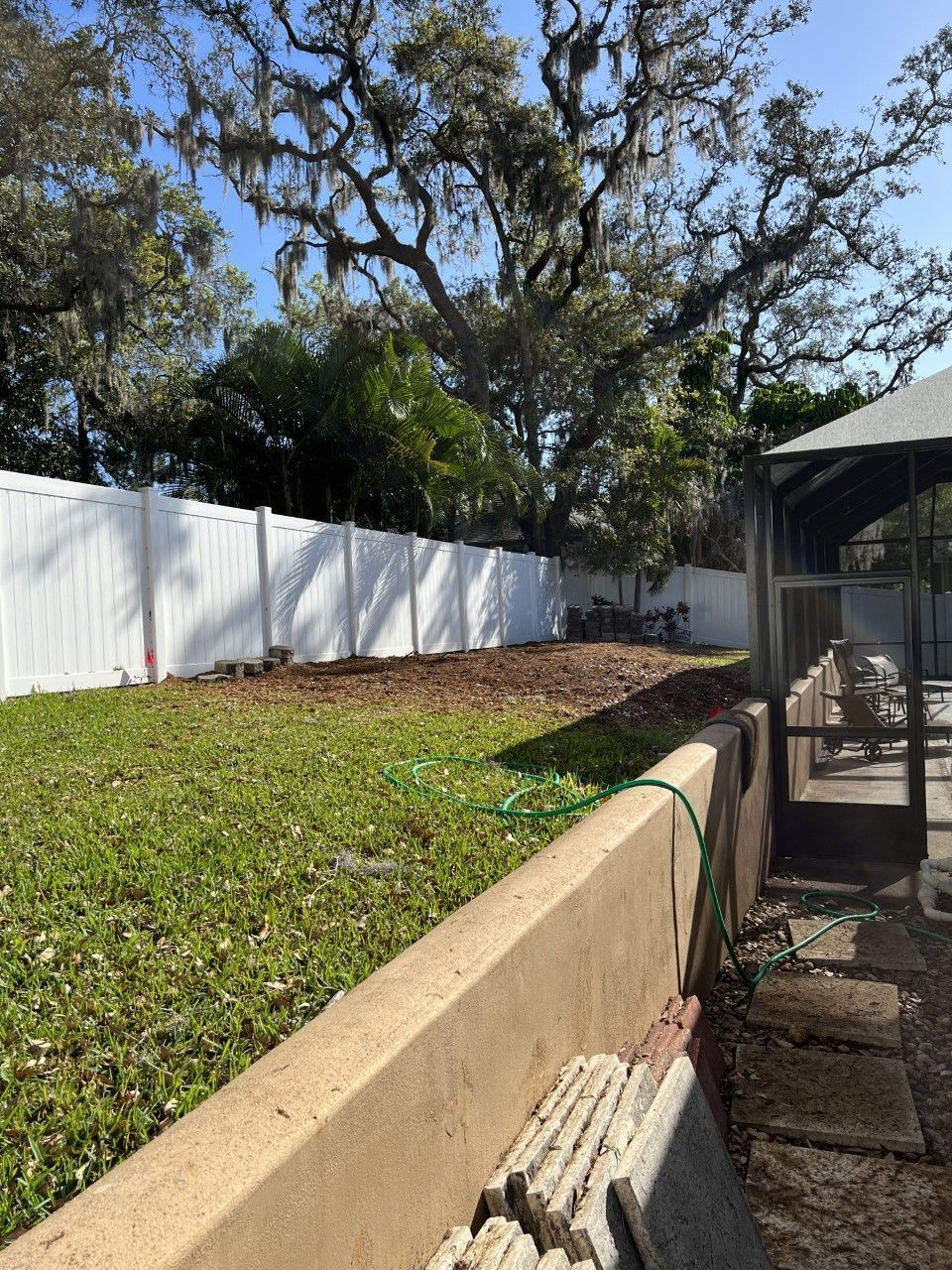 Backyard with white fence, grassy slope, retaining wall, and screened patio beside trees