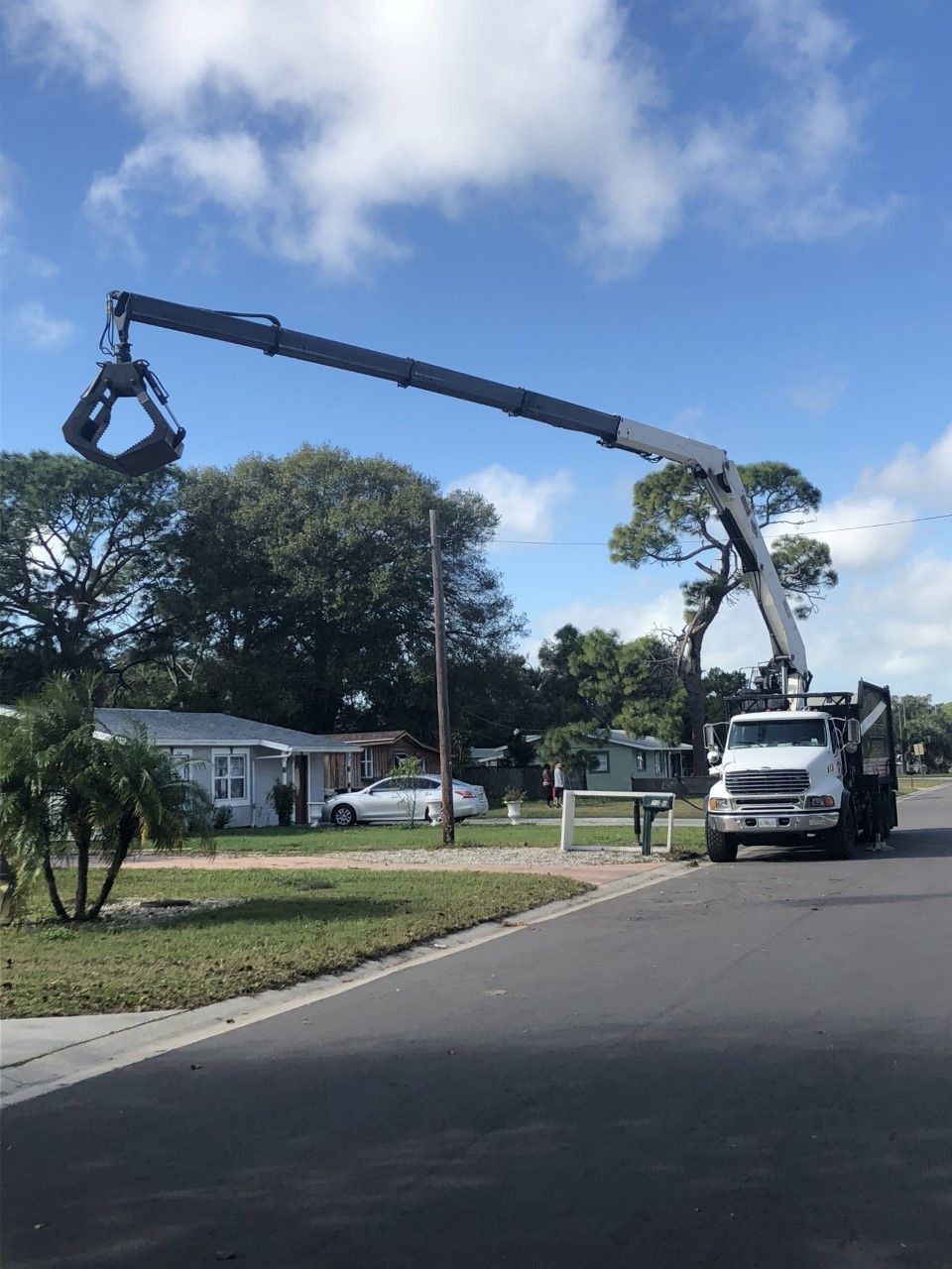 Truck-mounted crane lifting a broken tree branch over a suburban street under a blue sky