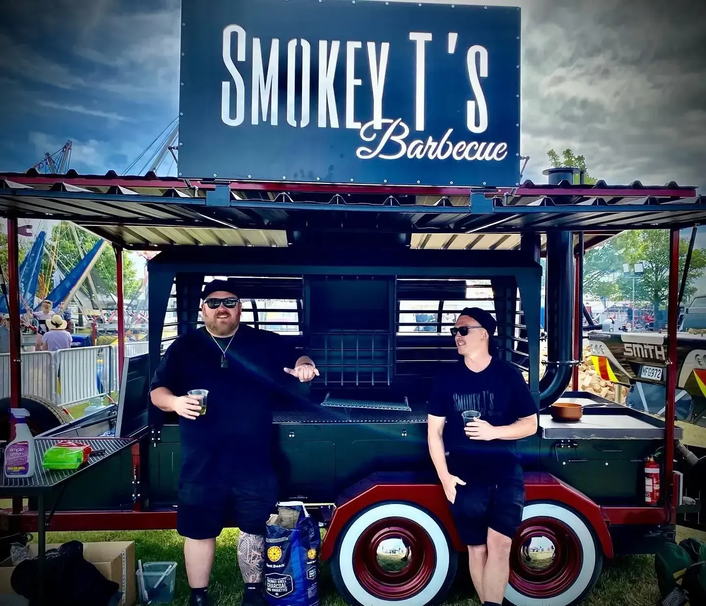 Two men are standing in front of a smokey t 's barbecue trailer.