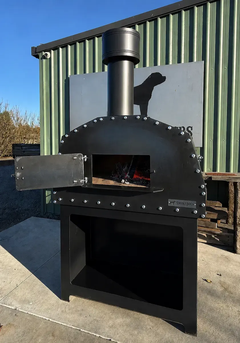 Black outdoor wood-fired oven with chimney and open front, set against a corrugated metal wall.