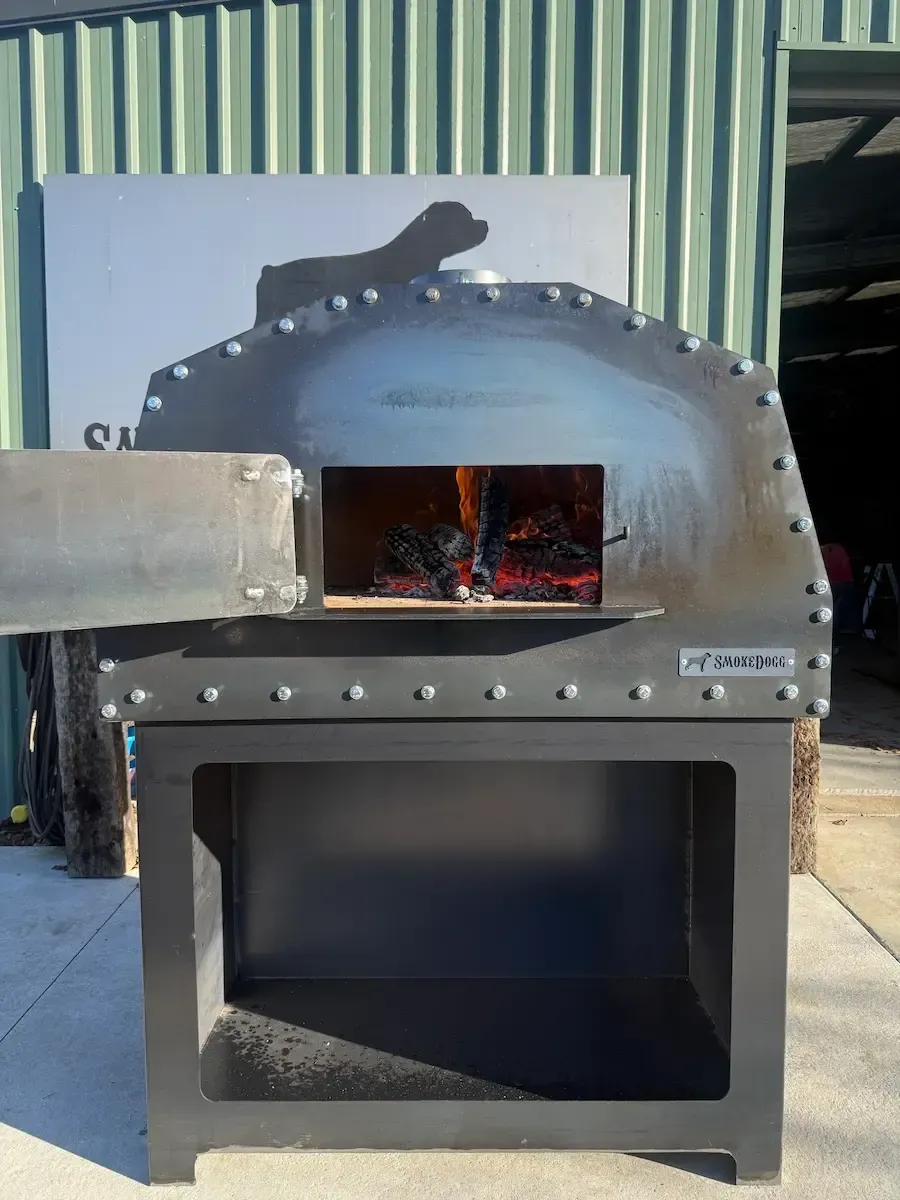 Stainless steel wood-fired oven with flames visible in the upper chamber, outdoors against a corrugated wall