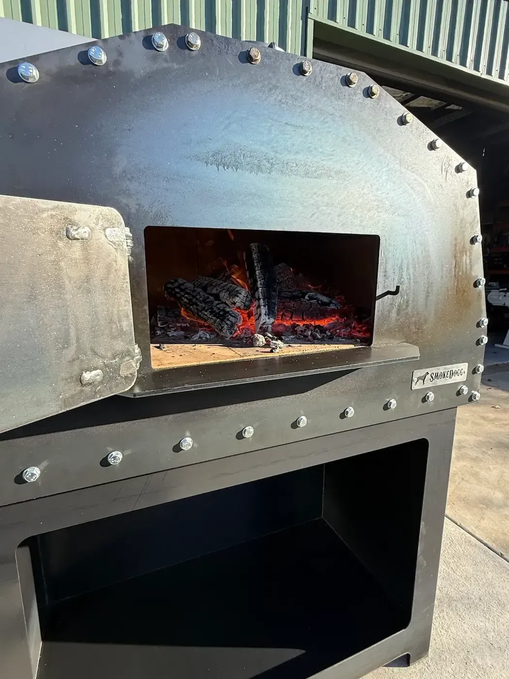Black metal wood-fired oven with glowing embers inside, outdoors on a patio