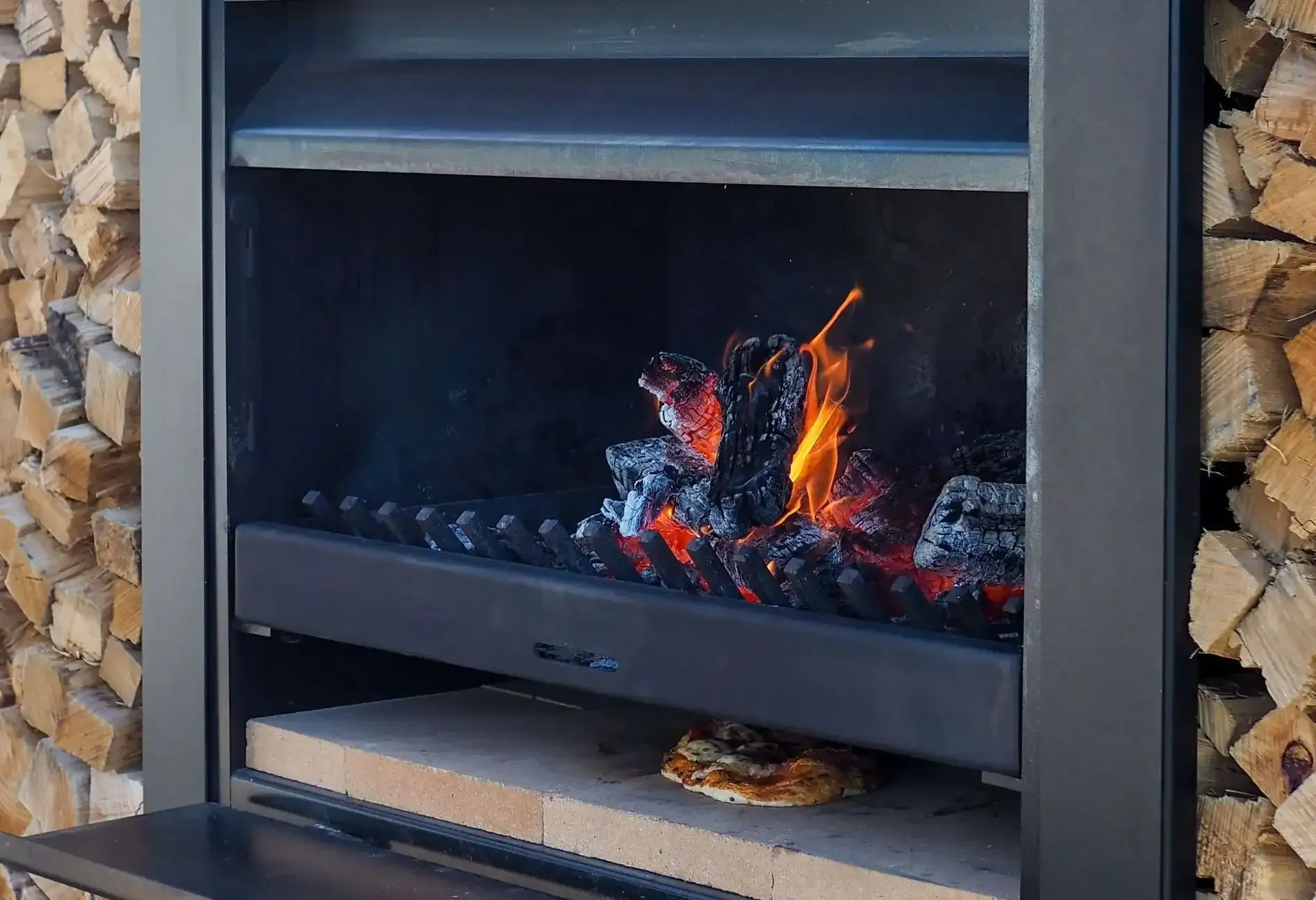 A pizza is cooking in a fireplace next to a pile of wood.