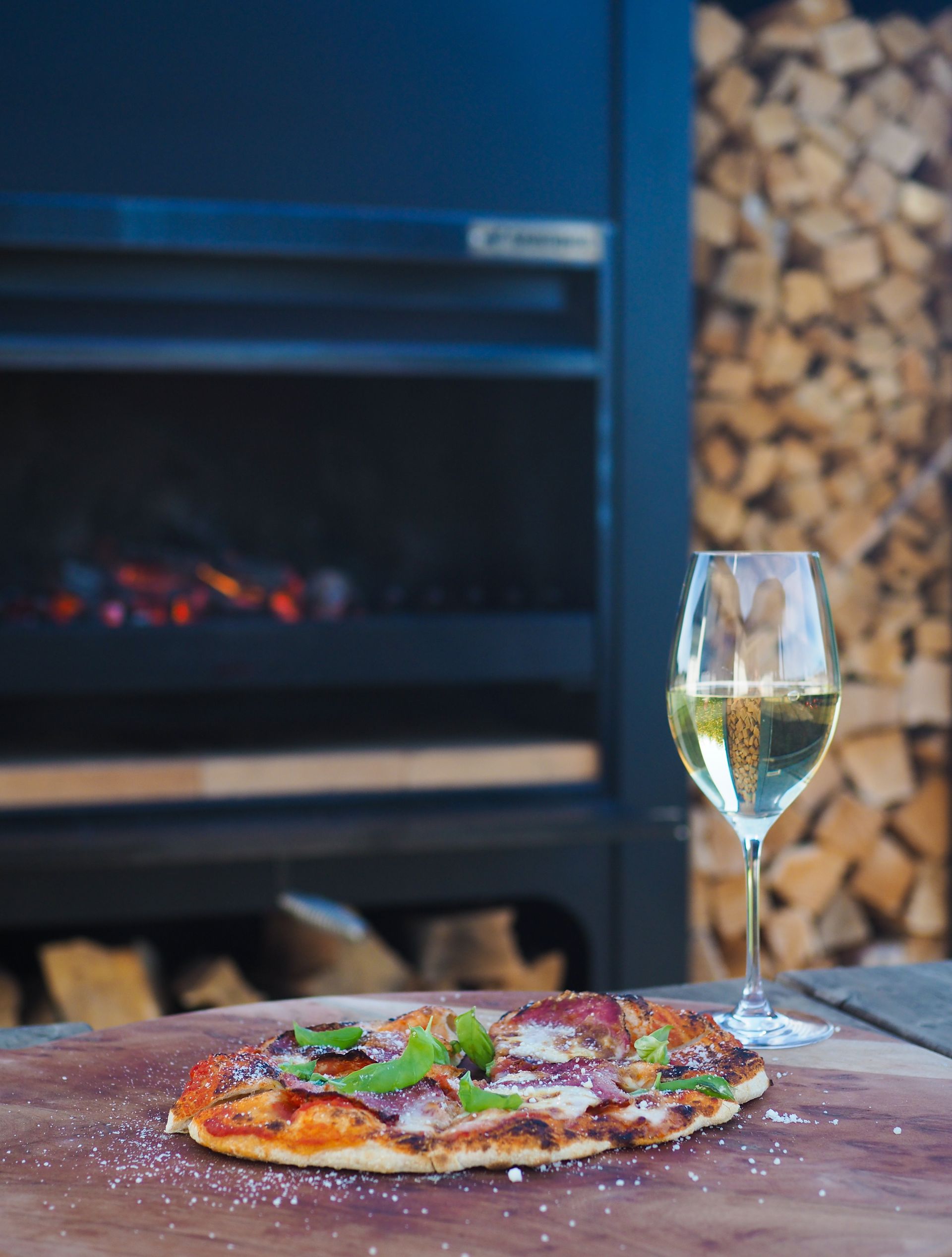 A pizza and a glass of wine are on a wooden table in front of a fireplace.