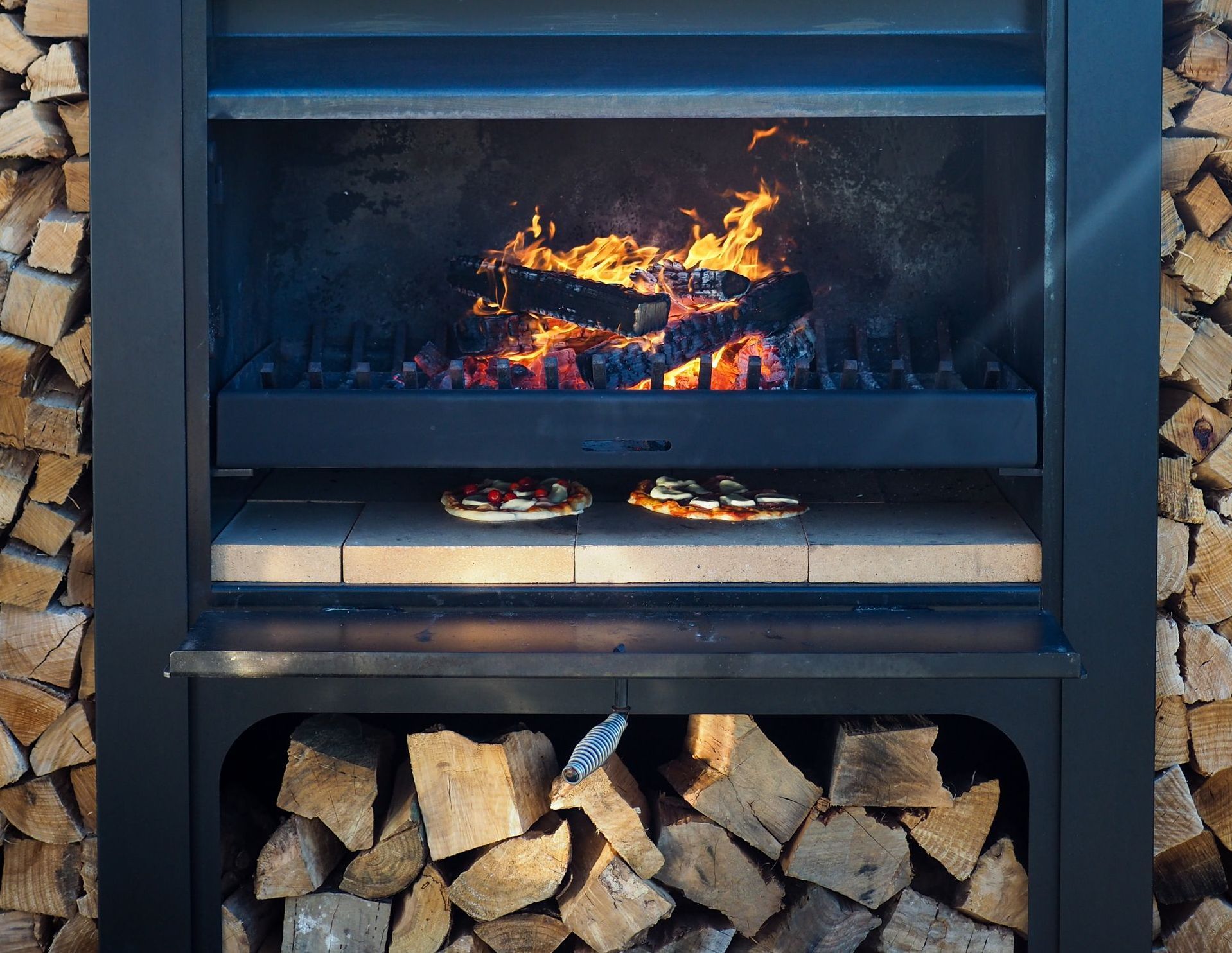 Two pizzas are cooking in a fireplace next to a pile of wood.