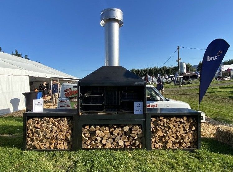 A large grill is sitting in the grass next to a pile of wood.