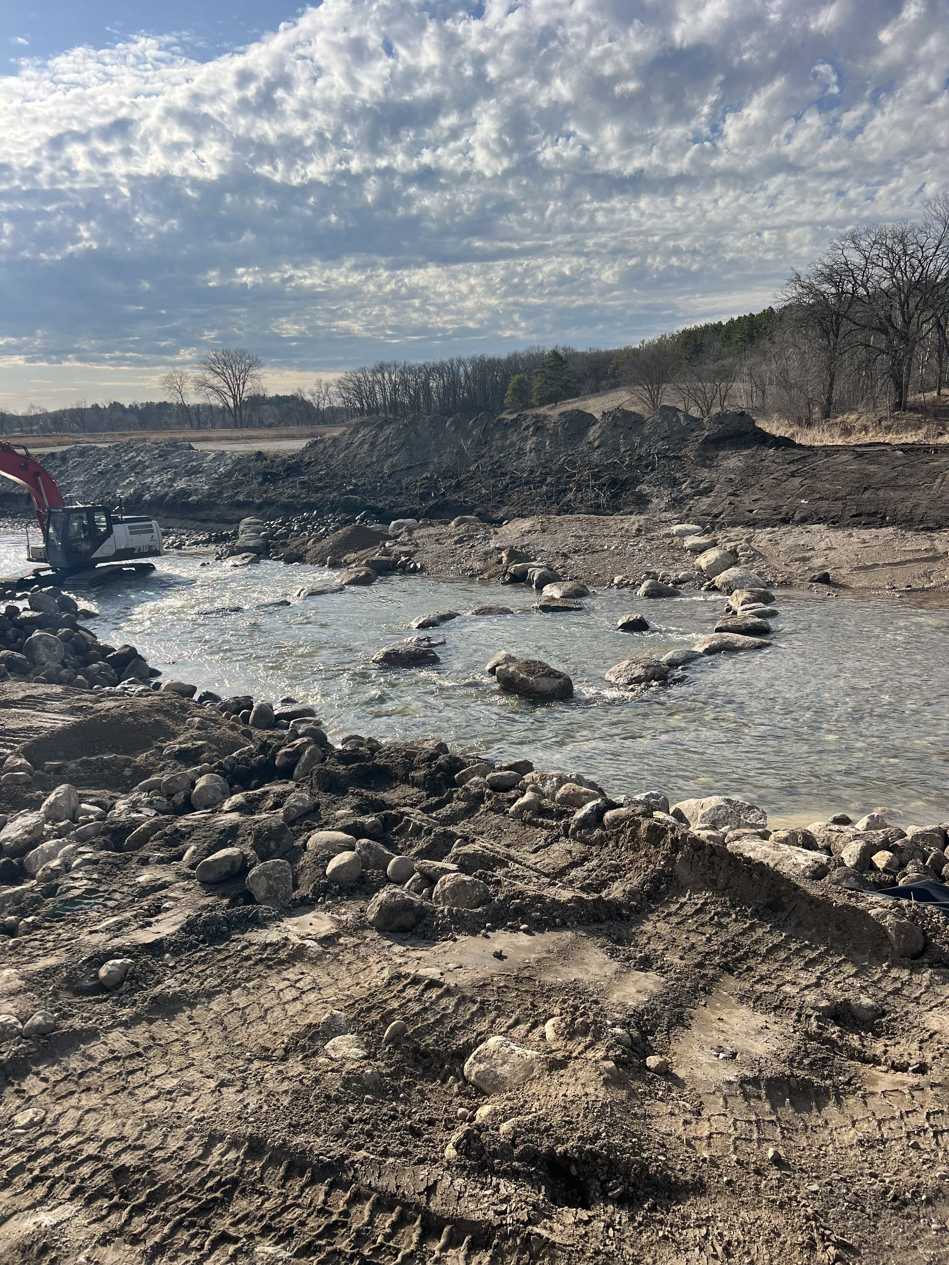Rocky riverbank with shallow water flowing under a cloudy sky and a red excavator at left.