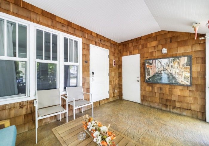 Empty sunroom with wood-shingle walls, white chairs, windows, and a floral painting on the wall