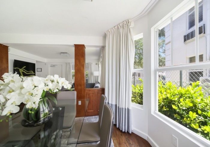Bright dining room with white flowers, glass table, and large windows overlooking greenery