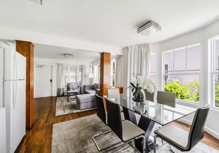 Bright dining room with glass table, black chairs, hardwood floor, and large windows with plants