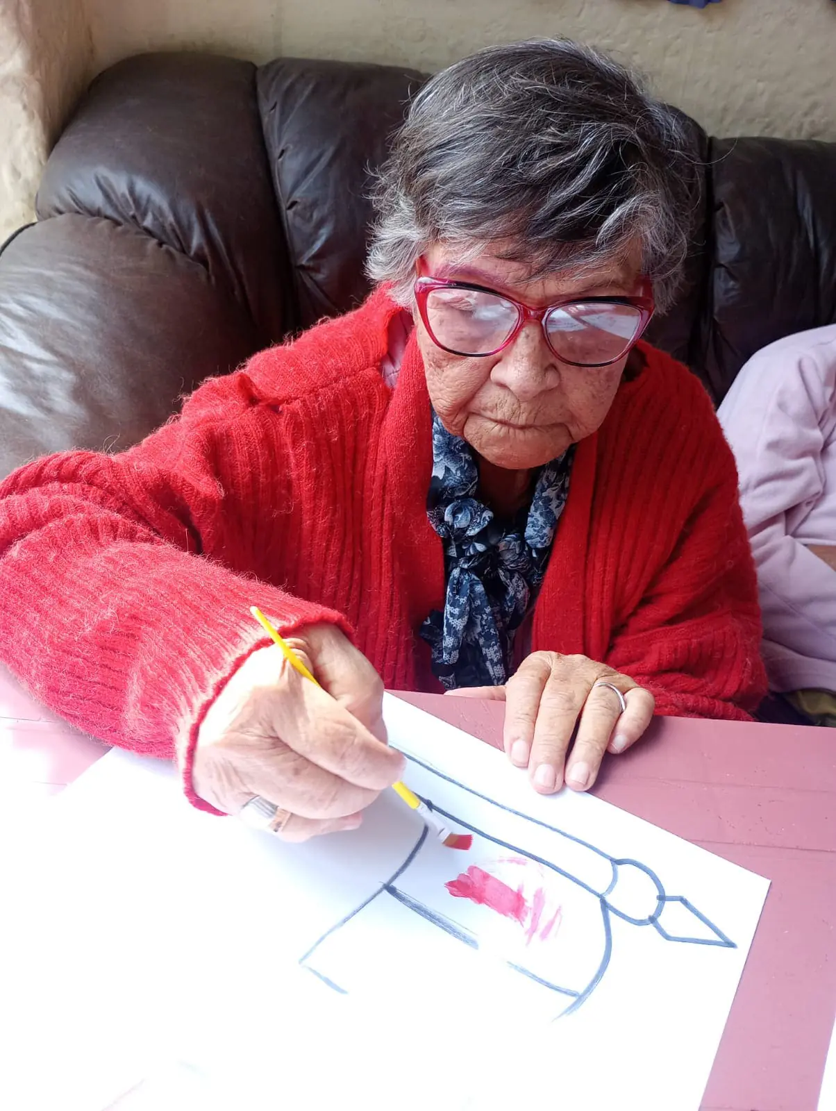 Mujer mayor con gafas dibujando en papel, con suéter rojo. Interior.