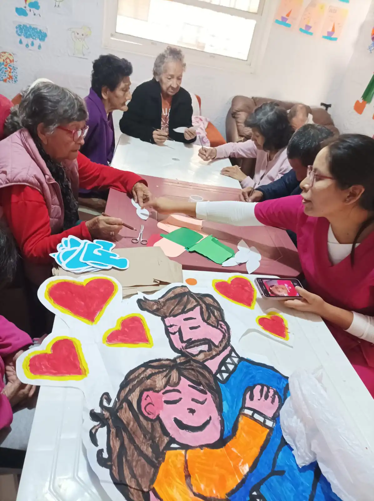 Personas mayores elaboran arte para el Día de San Valentín con un cuidador en una mesa; hay corazones coloridos y una ilustración de pareja.
