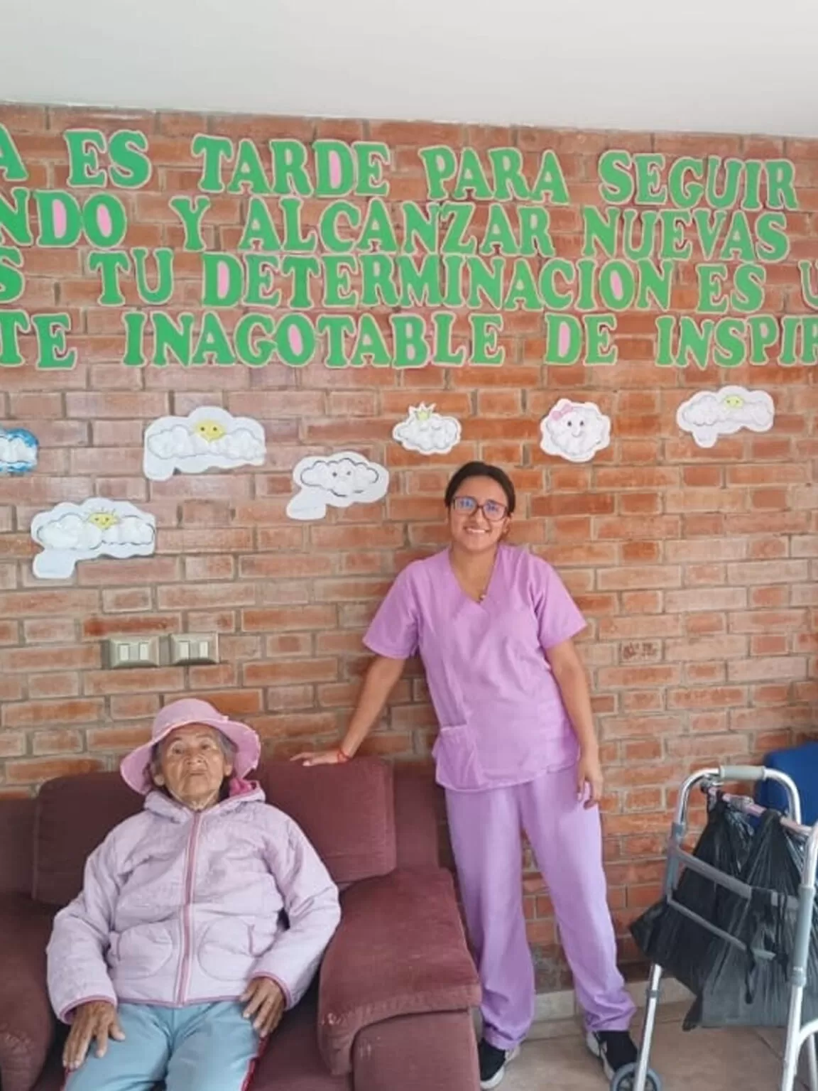 Una mujer con uniforme médico sonríe junto a una mujer mayor con ropa rosa, con una pared de ladrillos con escritura detrás de ellas.