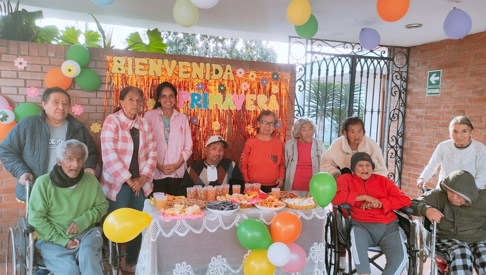 Grupo de personas, en su mayoría personas mayores, celebrando un evento primaveral con comida, globos y una pancarta de