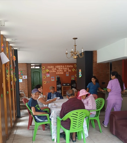 Un grupo de personas sentadas en una mesa, atendido por dos trabajadores de la salud en una sala con decoraciones coloridas.