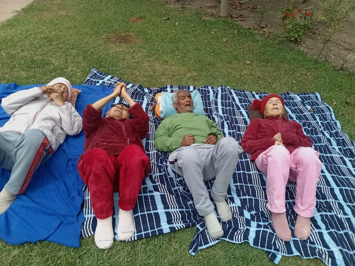 Cuatro personas mayores descansan sobre mantas en una zona de césped al aire libre. Llevan ropa de abrigo y disfrutan del sol.