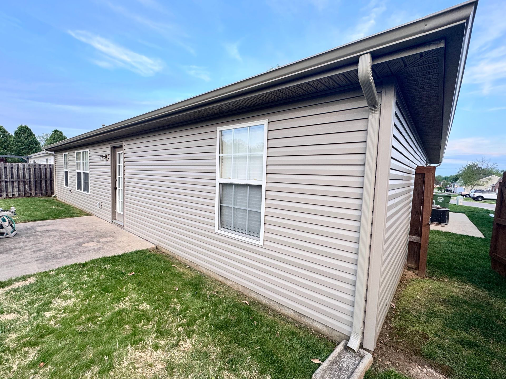 Single-story beige vinyl-sided house exterior with white trim and a small lawn and patio.