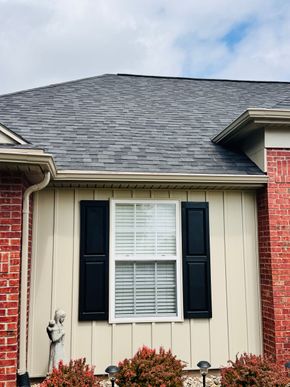 House exterior with tan siding, red brick, black shutters, and a gray shingled roof