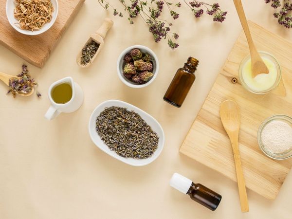 Ingredientes a base de hierbas y aceites para el cuidado de la piel, con cucharas de madera sobre una superficie beige.