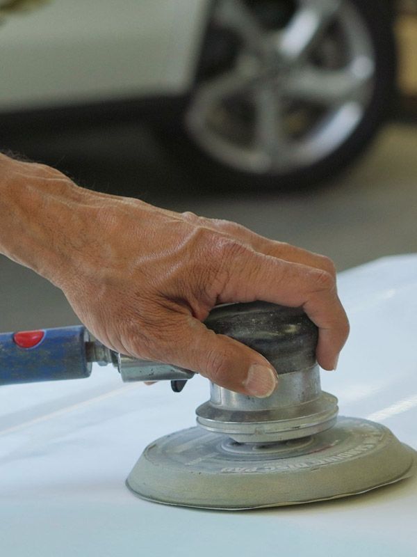 Man's hand holding an orbital sander and sanding a car hood.