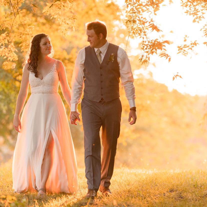 Bride and groom walking hand-in-hand through a golden-lit outdoor setting, surrounded by soft sunlig