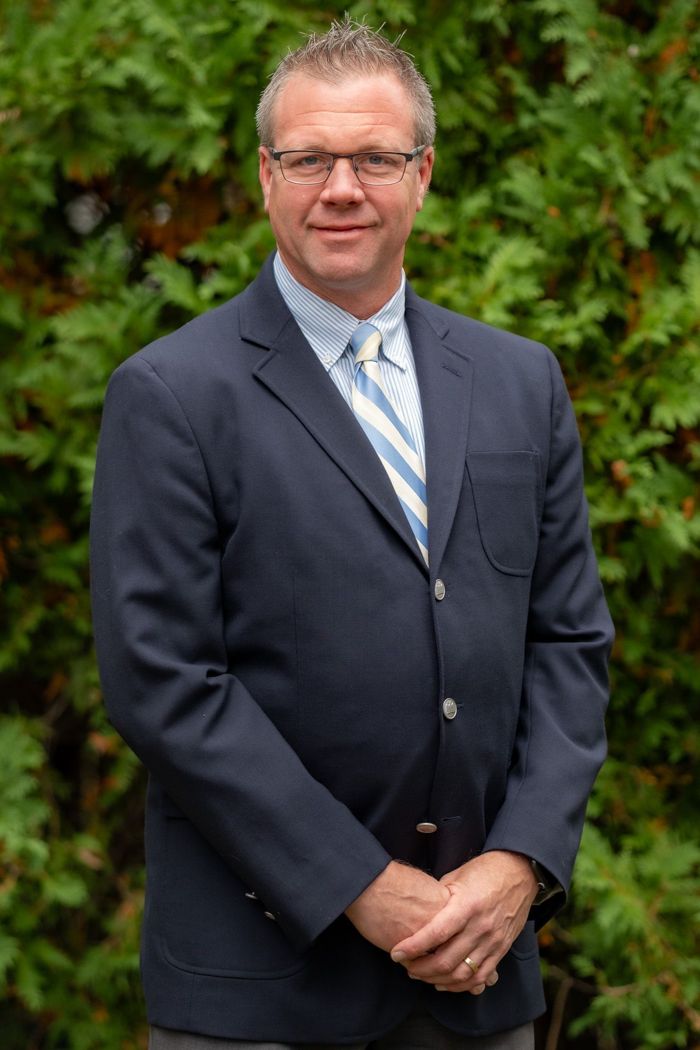 Man in a dark blazer and striped tie stands outdoors in front of green hedges, hands clasped.