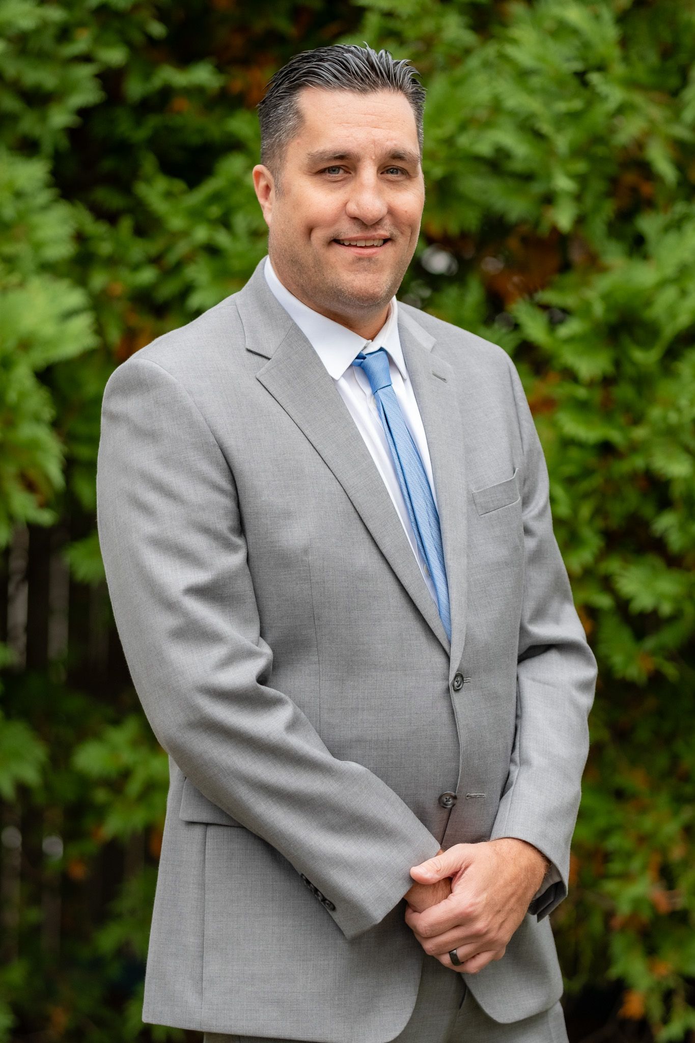 Man in a gray suit and blue tie standing outdoors in front of green foliage, hands clasped.