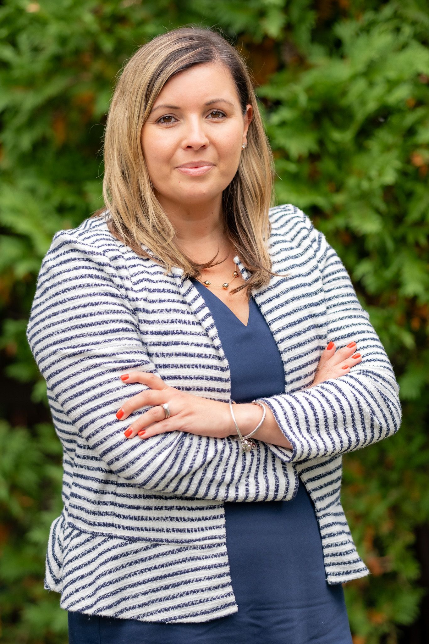 Person standing outdoors with arms crossed, wearing a striped blazer against green foliage.