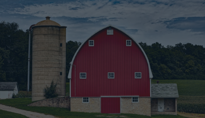 Red barn with white trim beside a tall silo in a rural field at dusk.