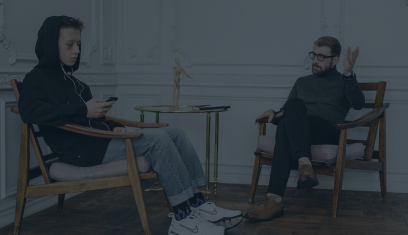 Two people sit facing each other in a minimalist room, talking across a small table.