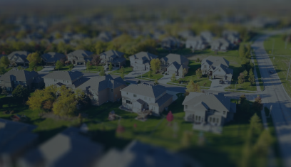 Aerial view of a suburban neighborhood with rows of houses, trees, and a road along the right side.