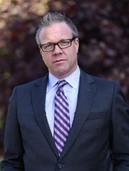 Man in a dark suit and striped tie standing outdoors against a blurred background