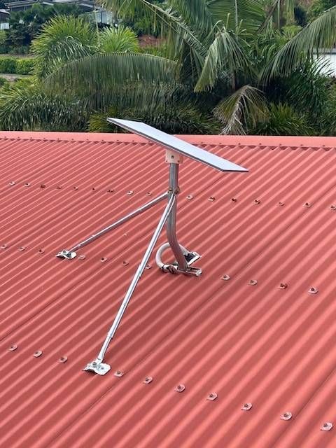 A Starlink internet dish is mounted on a red corrugated metal roof, angled upwards, against a background of green trees.