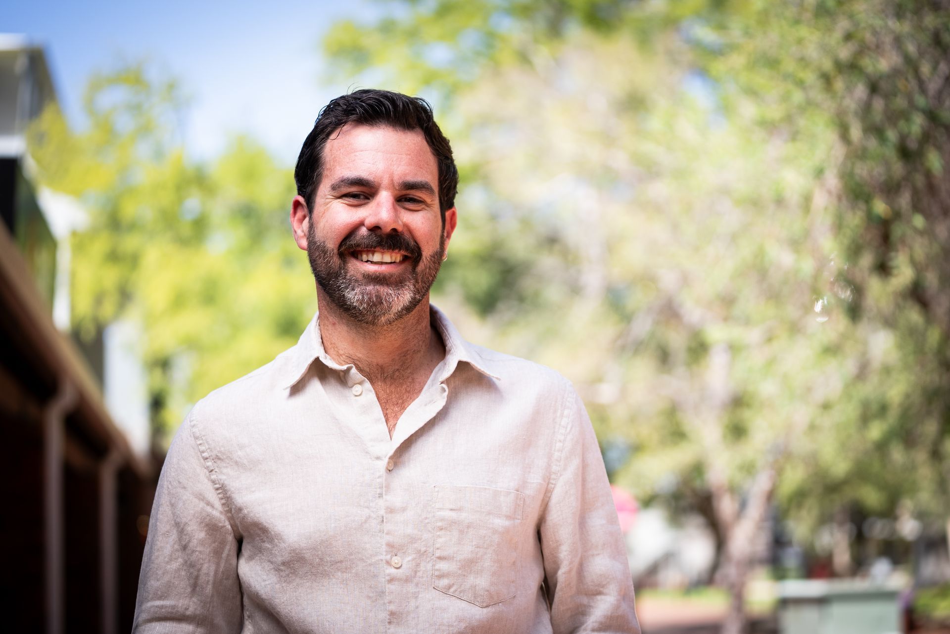 Man with a beard smiles, wearing a light-colored button-up shirt, outdoors in a green space.