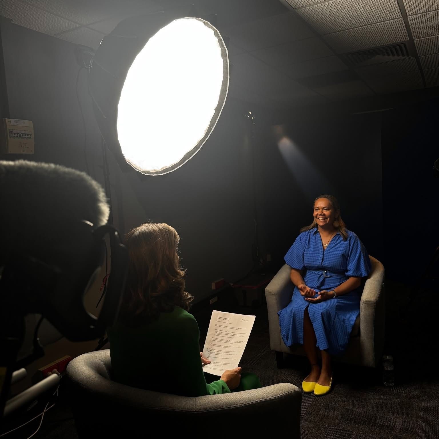 Woman being interviewed, seated in a studio. She wears a blue dress and smiles, facing the interviewer holding notes.
