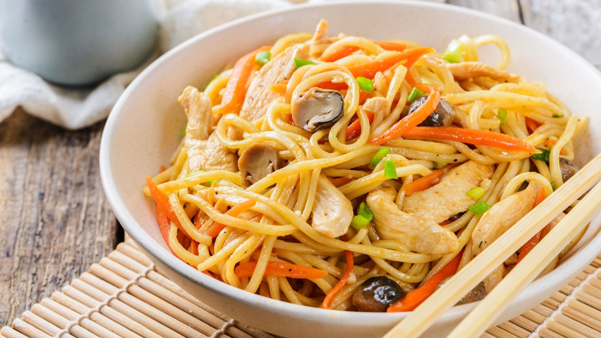A bowl of noodles with chicken , carrots , mushrooms and chopsticks on a wooden table.