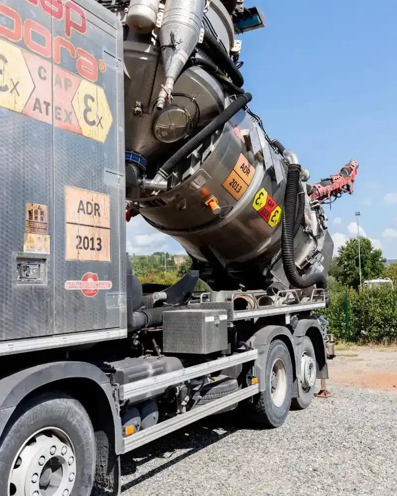 Un operaio in uniforme blu tiene in mano un grosso tubo collegato a un camion, probabilmente per lo smaltimento delle acque reflue, all'aperto.