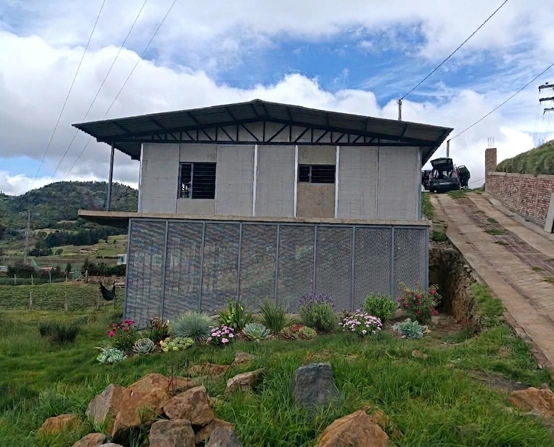 Pequeña casa rural con techo de metal en la ladera de una colina, rodeada de hierba, rocas y colinas a lo lejos.
