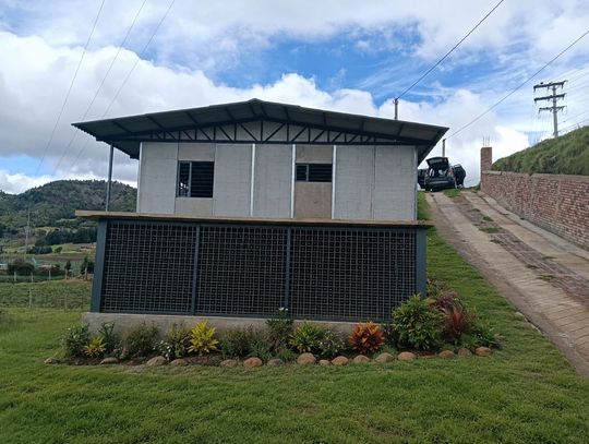 Pequeño edificio blanco en una ladera cubierta de hierba con un camino inclinado y cielo nublado.