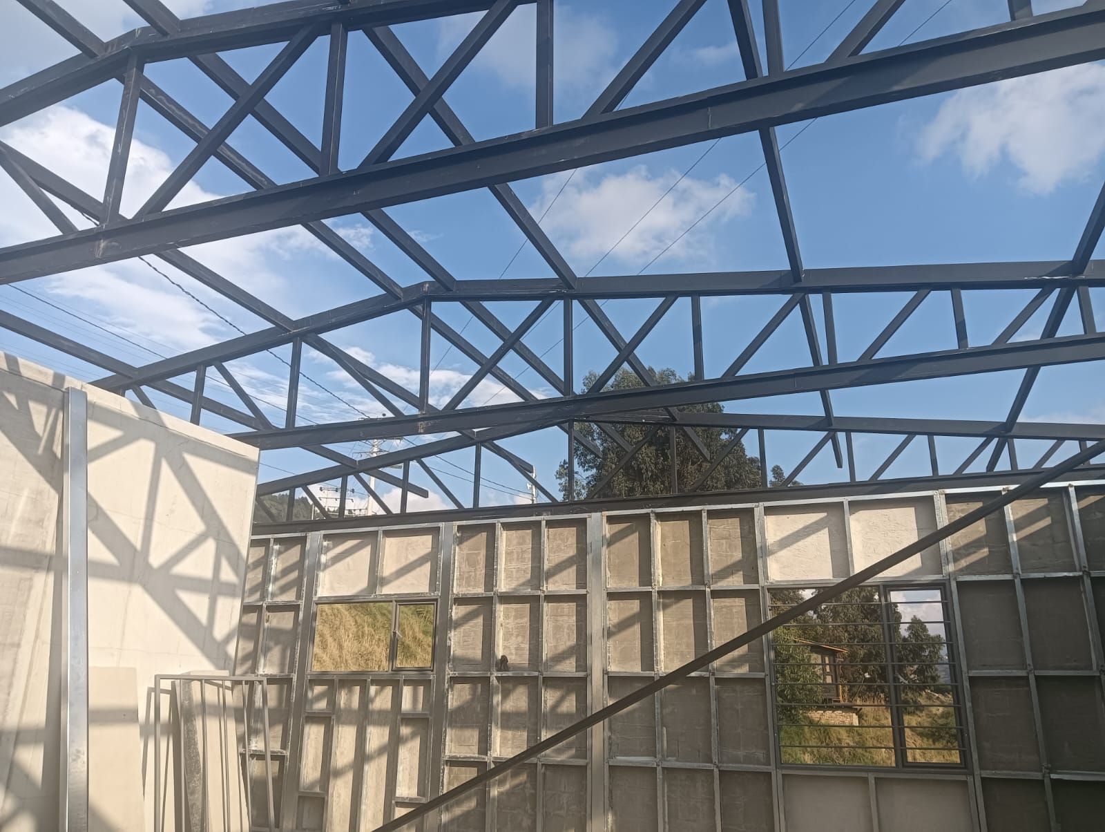 Estructura metálica parcialmente construida bajo un cielo azul, con paneles de pared y árboles visibles.
