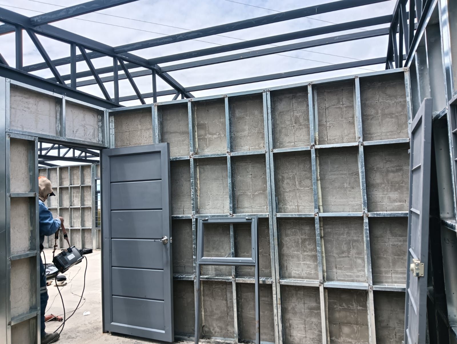 Interior de cobertizo sin terminar con estructura metálica, puerta abierta y montantes de pared a la vista en construcción.